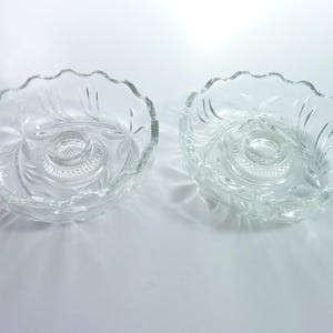 May include: Two clear glass candle holders featuring a scalloped rim and a central circular base with a textured, cross-hatched pattern. The glass surfaces are decorated with etched leaf-like motifs radiating from the center to the edges.