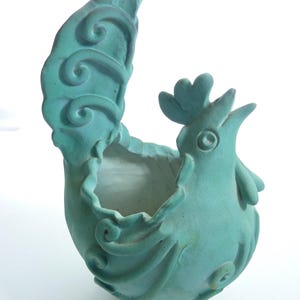 May include: A matte turquoise ceramic planter shaped like a stylized rooster. The vessel features a hollow center, a prominent upright tail with swirling embossed details, a comb on the head, and a wattle under the beak, all in a solid color.