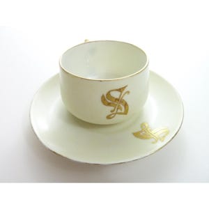 May include: Cream-colored teacup and saucer set with gold trim and a gold monogram. The teacup sits on the matching saucer. The teacup and saucer are made of porcelain.
