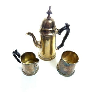 May include: A three-piece metal coffee service set featuring a tall coffee pot with a lid and black handle, a small creamer pitcher with a black handle, and a matching open cup. Each piece has a metallic finish with visible tarnish.