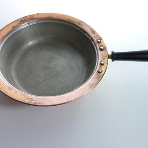 May include: A round copper pan with a silver-toned interior lining. The pan features a wide, flat copper rim and a single, smooth, black cylindrical handle attached to the side with three visible metal rivets. The copper surface shows signs of wear.