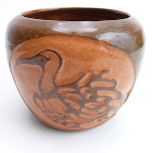May include: A round, hand-thrown ceramic bowl with a glossy dark brown rim and a lighter, reddish-brown body. The exterior features a debossed, stylized relief carving of a bird with wings tucked, showcasing a smooth, glazed finish throughout.
