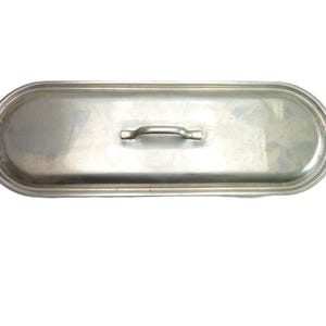 May include: An elongated, oval-shaped silver-toned metal lid featuring a central handle and two side handles. The surface has a smooth, reflective finish with a raised border around the perimeter, designed to fit a matching oblong cooking vessel.