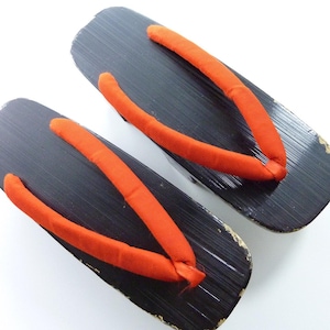 May include: A pair of traditional Japanese geta sandals featuring black lacquered wooden platforms with a textured grain finish and bright orange fabric thong straps. The wooden bases show minor wear along the edges, and the straps are secured.