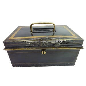 May include: A rectangular black metal cash box featuring a gold-toned handle on the lid, a central keyhole on the front, and decorative gold-colored border lines. The exterior finish shows signs of wear with visible paint chipping along the edges.
