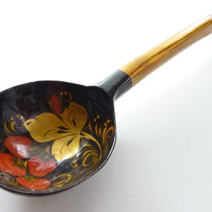 May include: A wooden ladle with a glossy black finish, featuring a hand-painted floral design in gold, red, and orange inside the bowl. The long handle transitions from black to a natural wood tone, ending with a curved red tip for a secure grip.