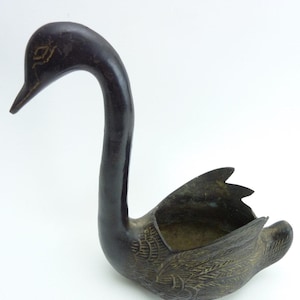 May include: A dark brown metal swan-shaped bowl with a long, curved neck and a hollow body. The exterior features etched feather details on the wings and a simple eye design on the head, with a metallic gold-toned finish visible in the crevices.