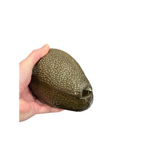 May include: A green avocado-shaped container with a textured surface. The container is held in a hand and has a small opening on one end. The container is made of a hard material.