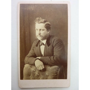 May include: A black and white portrait photograph of a man wearing a suit and tie. He is sitting in a chair with his arms crossed. The photo is in a vintage style.