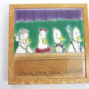 May include: A square ceramic tile in a light wood frame featuring a cartoon illustration of four white birds behind a wooden counter. The birds wear various accessories including hats, ties, and a bow tie. The text "DUCK, DUCK, DUCK, GOOSE" is written in black ink at the bottom.