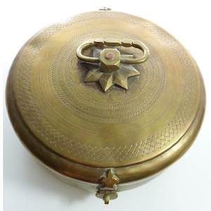 May include: A round brass container with a hinged lid featuring an engraved cross-hatch border and a central star-shaped ornament. A metal handle is attached to the top center, and a small latch mechanism is visible on the front edge of the lid.