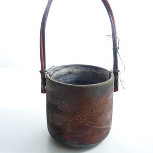 May include: A cylindrical ceramic vessel with a textured, dark brown and rust-colored exterior featuring subtle cross-hatched markings. The container includes a tall, arched handle made of thin, dark brown wooden strips secured with wire.