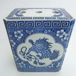 May include: A blue and white porcelain vase with a square shape and a decorative design. The vase features a detailed pattern of a mythical creature, floral motifs, and geometric borders. The top has three holes.