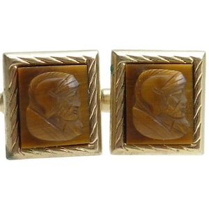 May include: A pair of vintage gold-toned cufflinks. Each cufflink features a square frame with a textured border, surrounding a rectangular, brown stone with a carved profile of a person. The cufflinks are connected by a gold-toned bar.