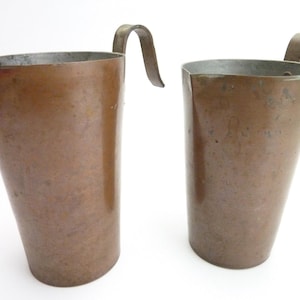 May include: Two copper pitchers with a weathered, metallic finish and a tapered cylindrical shape. Each vessel features a wide, open top with a distinct pouring spout and a curved metal handle attached to the side, showing signs of oxidation.