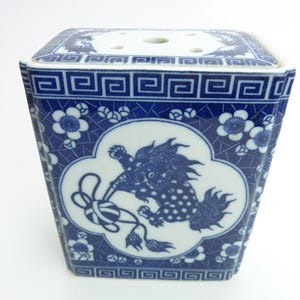 May include: A blue and white ceramic flower frog with a square shape. The sides feature a repeating floral and geometric pattern. A central panel depicts a mythical creature. The top has several holes for arranging flowers.