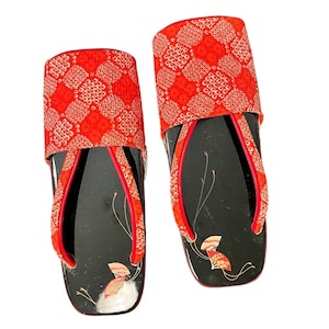 May include: A pair of traditional Japanese zori sandals featuring black lacquered footbeds with delicate fan illustrations. The wide, padded straps are covered in a vibrant red fabric with a white geometric shibori-style diamond pattern.