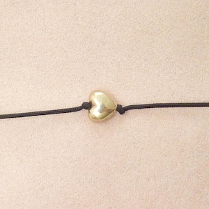 May include: A gold-colored heart-shaped charm centered on a thin black cord. The heart has a smooth, reflective metallic surface and is secured in place by small knots on either side of the charm along the length of the black string.