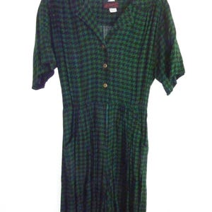 May include: A short-sleeve jumpsuit featuring a dark green and black houndstooth pattern. The garment has a collared neckline, a button-down front closure, and an elasticized waist with two side pockets. The fabric has a soft, draped appearance.