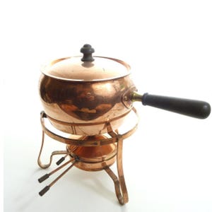 May include: A copper fondue pot with a matching lid and a long black handle. The pot rests on a copper stand that features a small burner underneath. The metal surfaces have a polished finish, and the handle is cylindrical and dark in color.