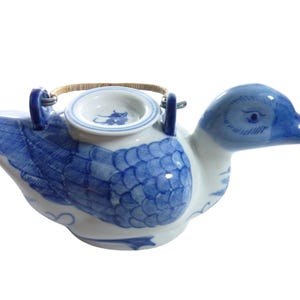 May include: A ceramic duck-shaped teapot with a blue and white design. The teapot features a detailed blue feather pattern on its body and a blue head. It has a small, round opening on top and a woven handle.
