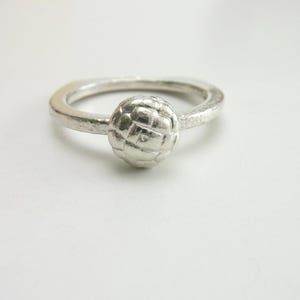 May include: A silver ring featuring a textured band and a central spherical ornament. The sphere has a grid-like pattern etched into its surface, and the entire piece has a matte, metallic finish with visible handcrafted detailing throughout.