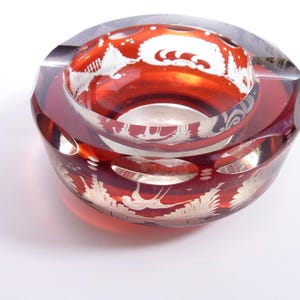 May include: A round, red glass ashtray with etched white floral and bird designs. The ashtray has a thick, clear rim and several oval-shaped indentations for resting cigarettes. The interior is a vibrant red color.