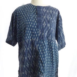 May include: A short-sleeved shirt in a deep indigo blue with a repeating light gray diamond ikat pattern. The shirt features a partial button-down placket at the center front, a round neckline, and a single chest pocket on the left side.