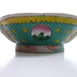 May include: A decorative ceramic bowl with a wide, shallow shape. The bowl features a teal green exterior with yellow and pink accents. The rim has a scalloped edge. The bowl's design includes floral and landscape motifs.