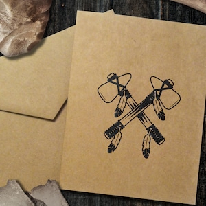 May include: Kraft paper greeting cards with a black illustration of crossed tomahawks and feathers. An envelope and a vintage photograph are also visible. A stone axe with a wooden handle is in the background.