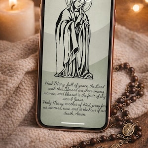 May include: Smartphone displaying a black and white illustration of the Virgin Mary with a halo, and the Hail Mary prayer. A rosary with a crucifix lies nearby. A lit candle is in the background.