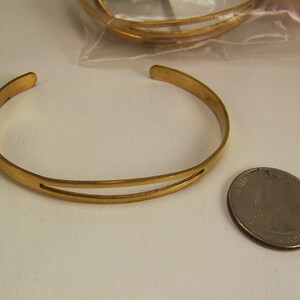 May include: A gold-colored metal cuff bracelet with a split design. The bracelet is open-ended and sits next to a quarter coin. Additional bracelets are visible in the background.