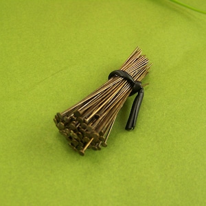 May include: A bundle of thin, metallic, bronze-colored head pins with flat, circular tops. The pins are gathered together and secured in the center by a small, black, flexible rubber band that holds the cluster in a tight, cylindrical shape.