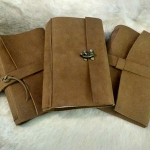 May include: Three brown leather-bound journals with clasps. The journal on the left has a leather strap and tie. The journal in the middle has a metal clasp. The journal on the right has a leather strap.