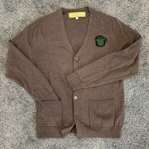 May include: A brown cardigan sweater with a green coffee cup patch on the front. The sweater has two pockets and a button closure.