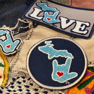 May include: Three embroidered patches featuring the state of Michigan in blue and white with a red heart. The patches are on a background of various fabrics. One patch says "LOVE" in white letters with a blue outline.