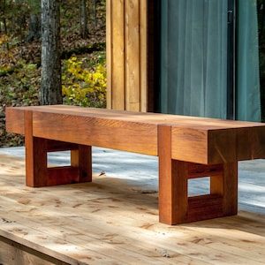 May include: A wooden bench with a natural reddish-brown finish. The bench has a simple design with a rectangular seat and square legs. The bench is sitting on a wooden deck.
