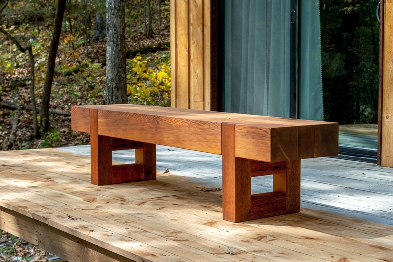 May include: A wooden bench with a natural reddish-brown finish. The bench has a simple design with a rectangular seat and square legs. The bench is sitting on a wooden deck.