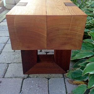 May include: A wooden bench with a natural finish and a dark brown base. The bench is made of two thick planks of wood joined together with metal brackets.