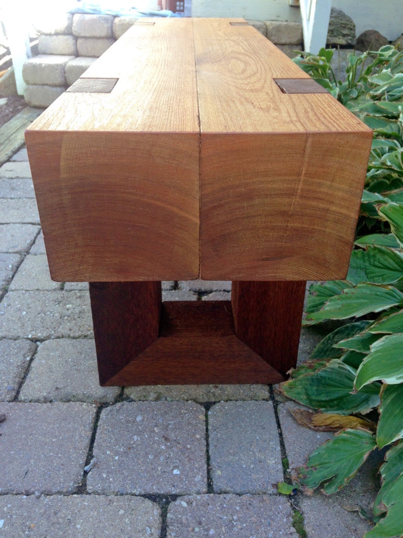 May include: A wooden bench with a natural finish and a dark brown base. The bench is made of two thick planks of wood joined together with metal brackets.