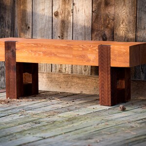 May include: A rustic wooden bench with a natural wood finish. The bench has a wide, flat seat and two sturdy legs made of a darker wood. The bench is sitting on a wooden deck.