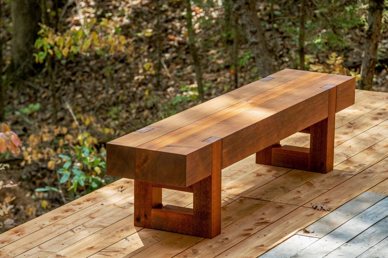 May include: A rustic wooden bench with a natural wood finish. The bench has a simple design with a rectangular seat and square legs. The bench is made of solid wood and has a natural wood grain.