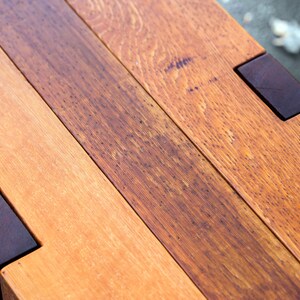 May include: Close-up of a wooden bench with a natural wood finish. The bench is made of multiple planks of wood, some of which are darker than others. The bench has a dark brown stain on the edges.