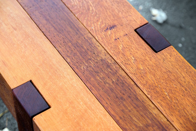 May include: Close-up of a wooden bench with a natural wood finish. The bench is made of multiple planks of wood, some of which are darker than others. The bench has a dark brown stain on the edges.