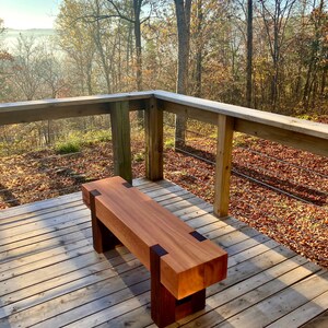 May include: A wooden bench with dark brown accents sits on a wooden deck overlooking a foggy forest. The bench is made of light brown wood and has a simple, rectangular design.