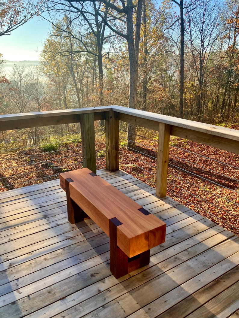 May include: A wooden bench with dark brown accents sits on a wooden deck overlooking a foggy forest. The bench is made of light brown wood and has a simple, rectangular design.