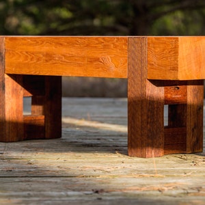 May include: A wooden bench with a natural finish and a simple design. The bench has a rectangular seat and four legs with a square shape. The legs are connected by a crossbar.