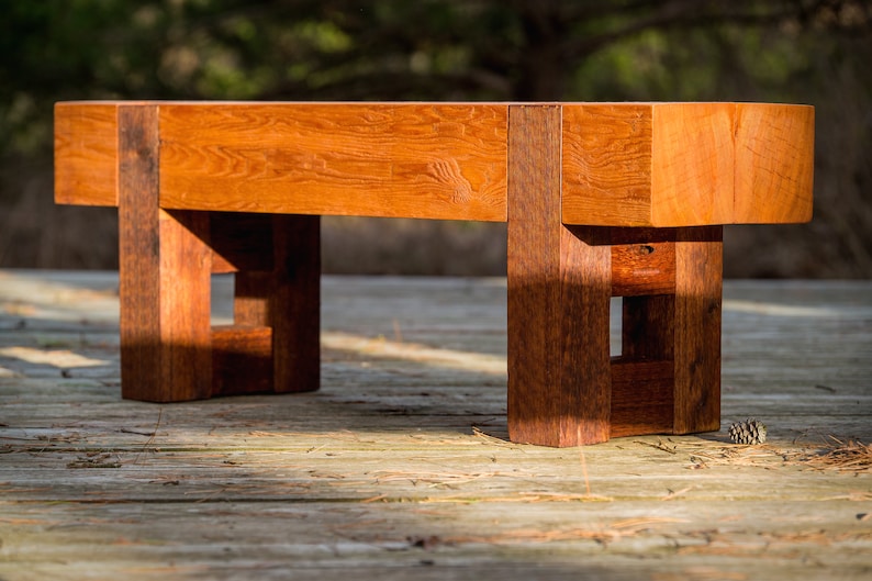 May include: A wooden bench with a natural finish and a simple design. The bench has a rectangular seat and four legs with a square shape. The legs are connected by a crossbar.