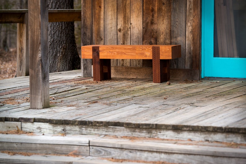 May include: A wooden bench with a natural finish sits on a wooden porch with a blue door in the background.