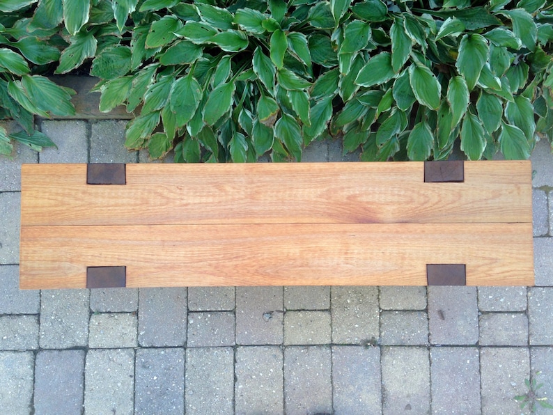 May include: A long wooden bench with four dark brown metal brackets on the top surface. The bench is made of two planks of wood and is sitting on a gray brick patio.